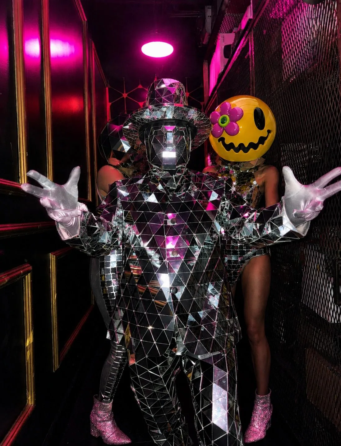 A dancer in a mirror-tile suit standing with a performer wearing a giant yellow smiley mask with a pink flower for an eye, in a pink lit corridor.