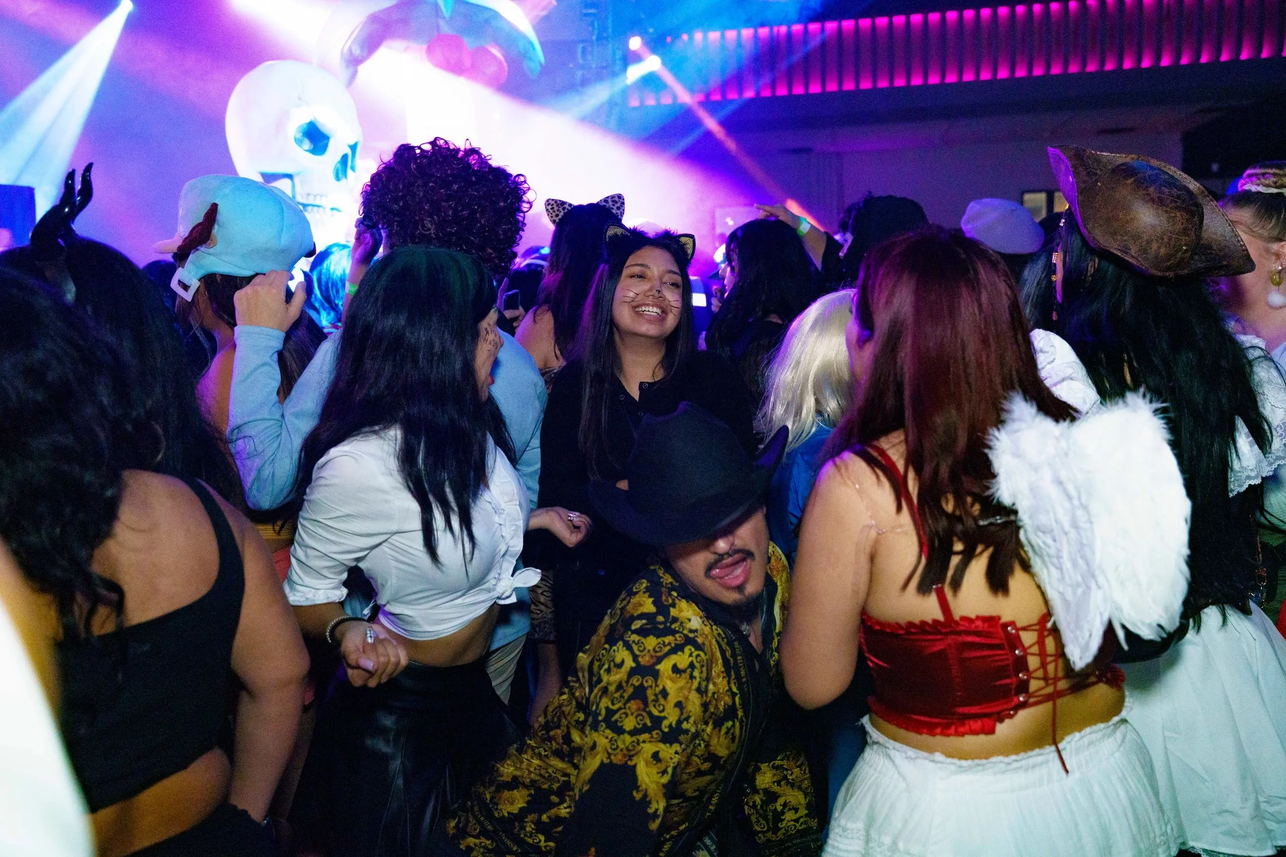 A Halloween crowd in costume with hands and phones in the air, glow bands on their wrists.