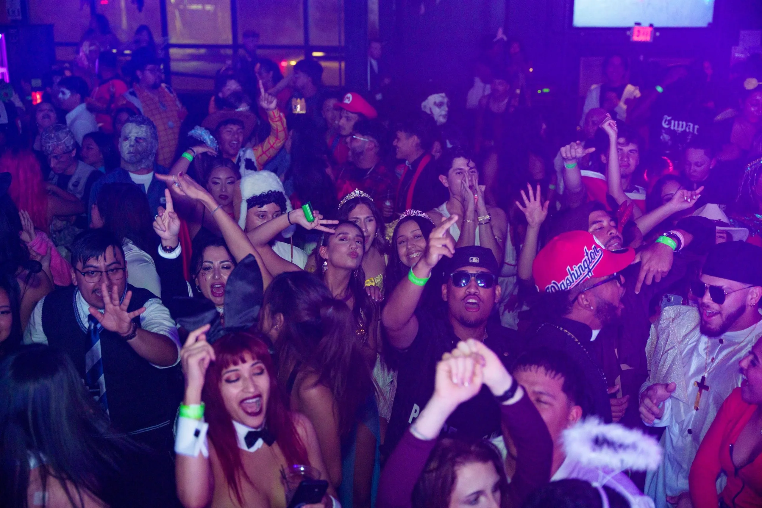 A dense Halloween crowd in costume with hands up, the room washed red and purple.