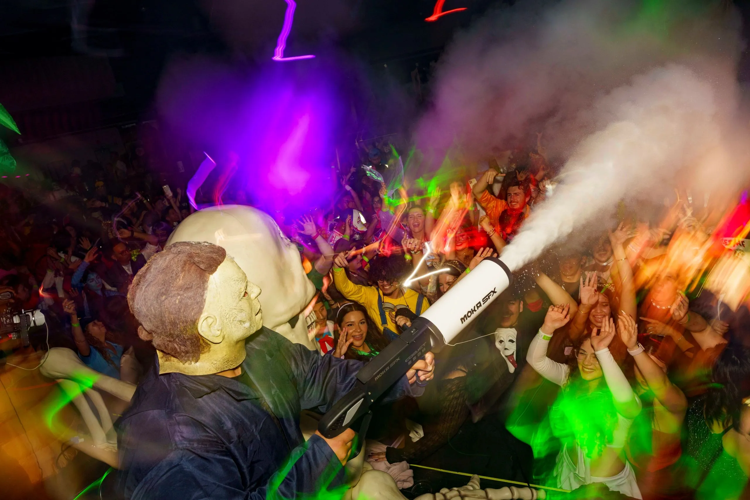 A crew member firing a CO2 cannon over the crowd in green and purple light, masked dancers behind him.
