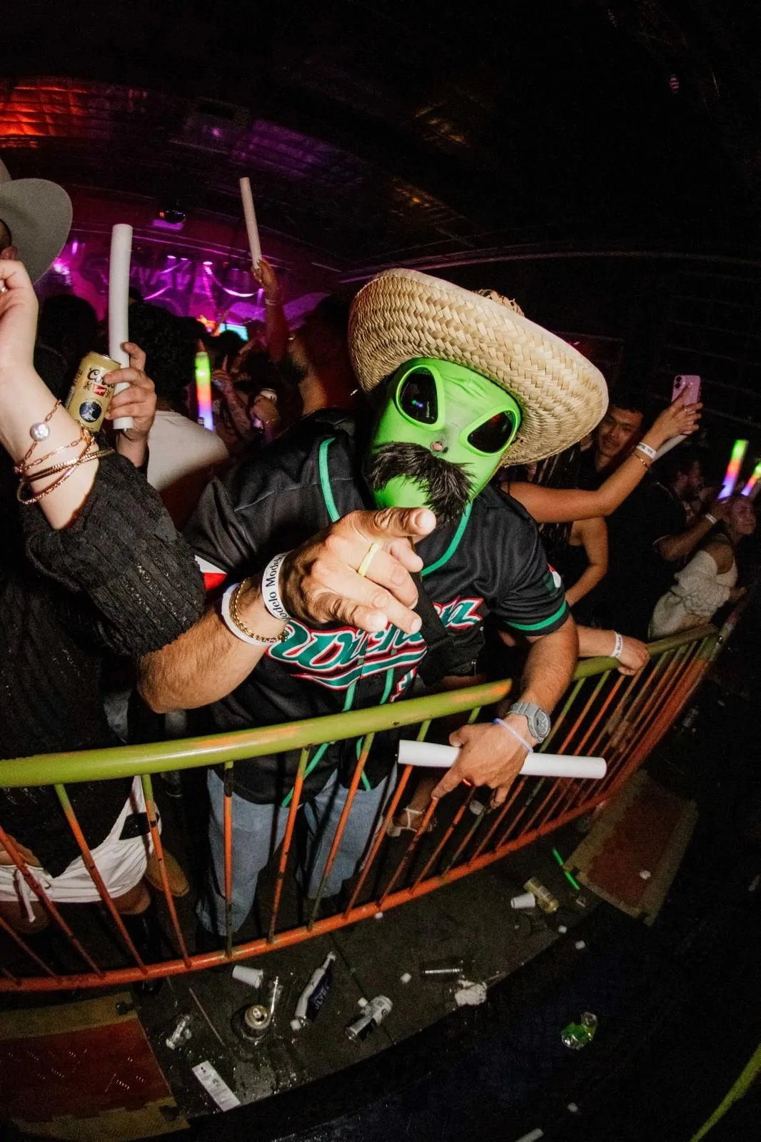 A guest at the rail in a green alien mask and a straw sombrero, holding a light stick.