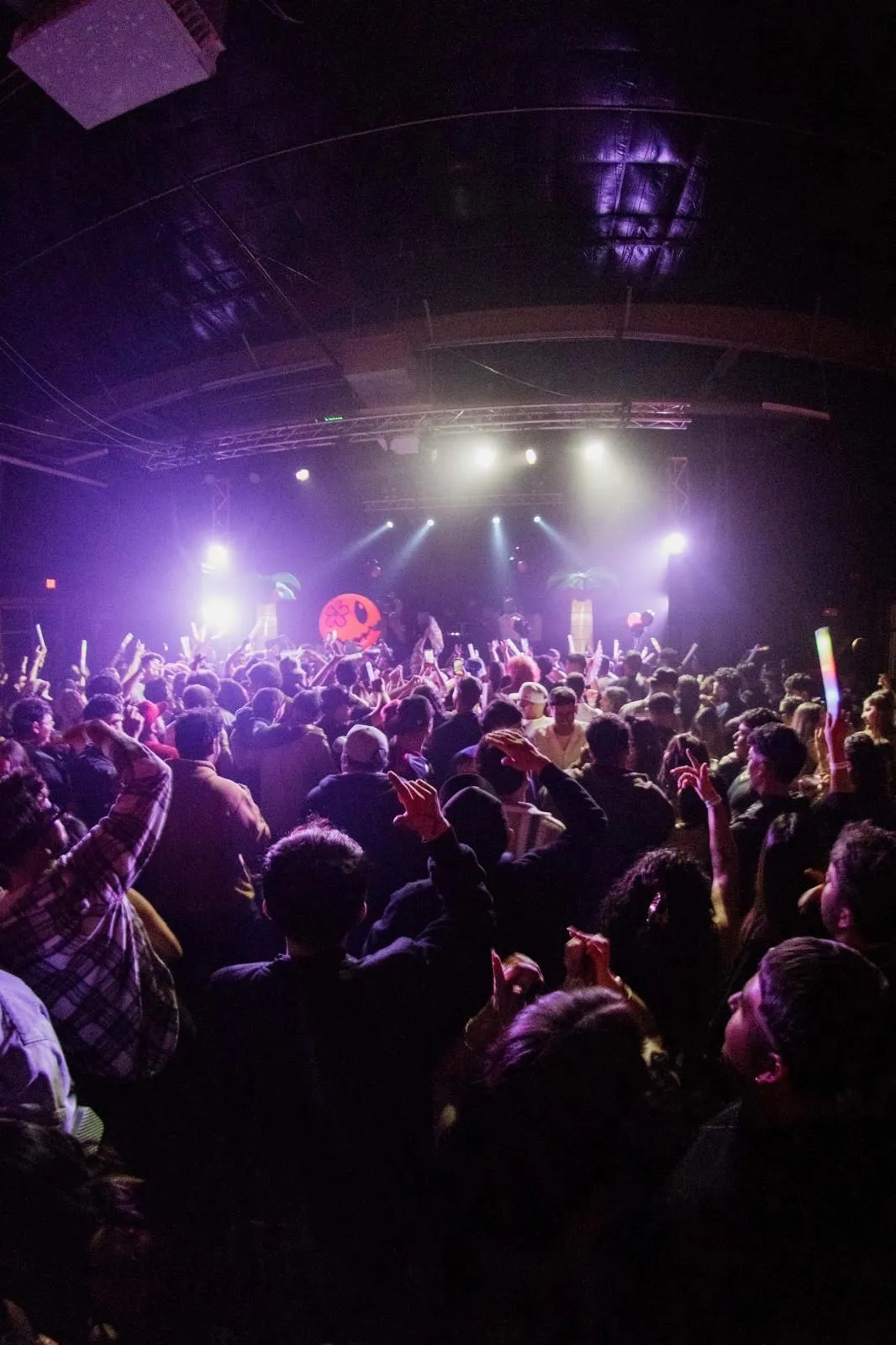 The back of a packed theatre floor looking toward the stage, hands and phones up under purple light.