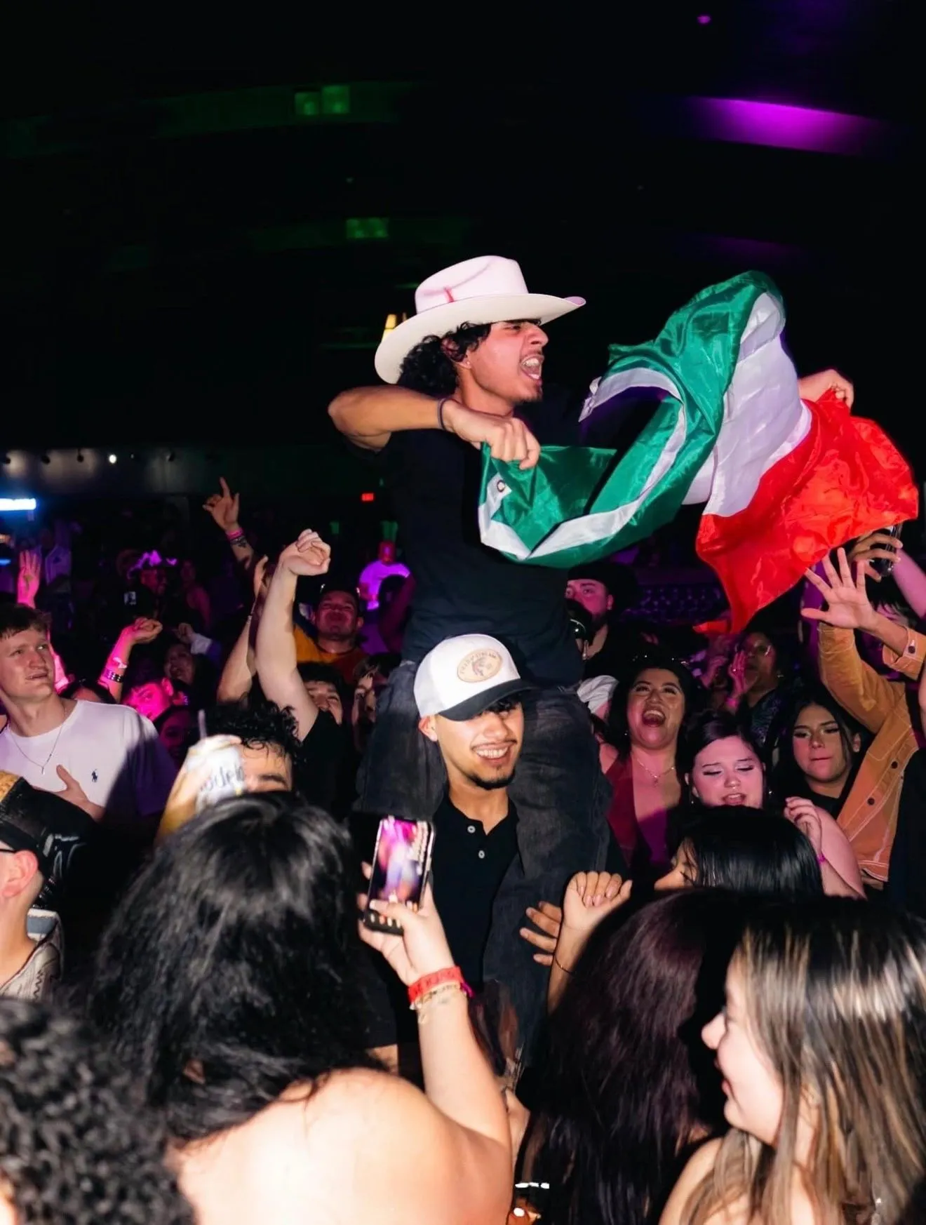 A man in a white cowboy hat sitting on a friend's shoulders, holding a Mexican flag open above the crowd.