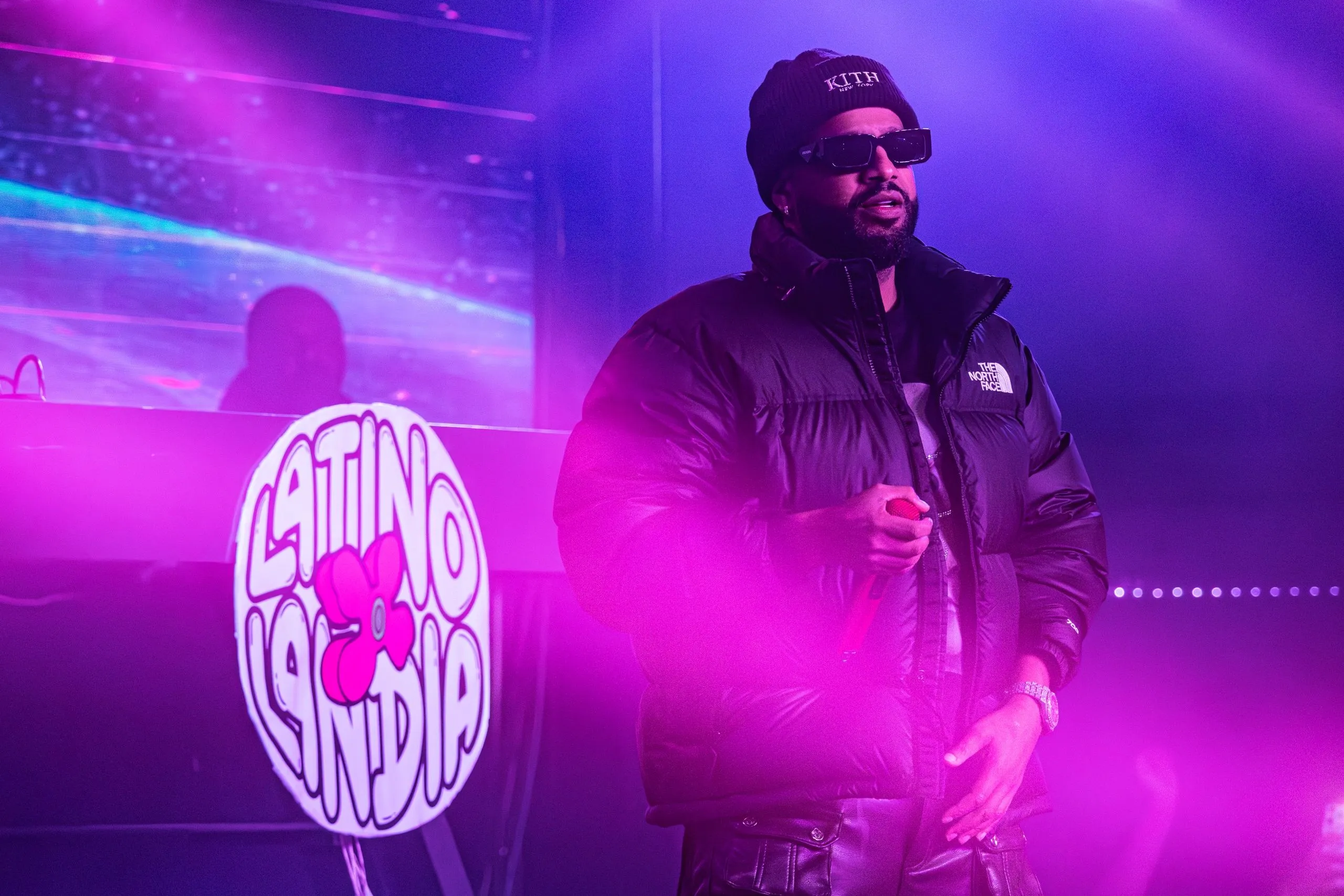 An artist on stage in a black puffer jacket beside a lit Latino Landia bubble logo backdrop under pink light.