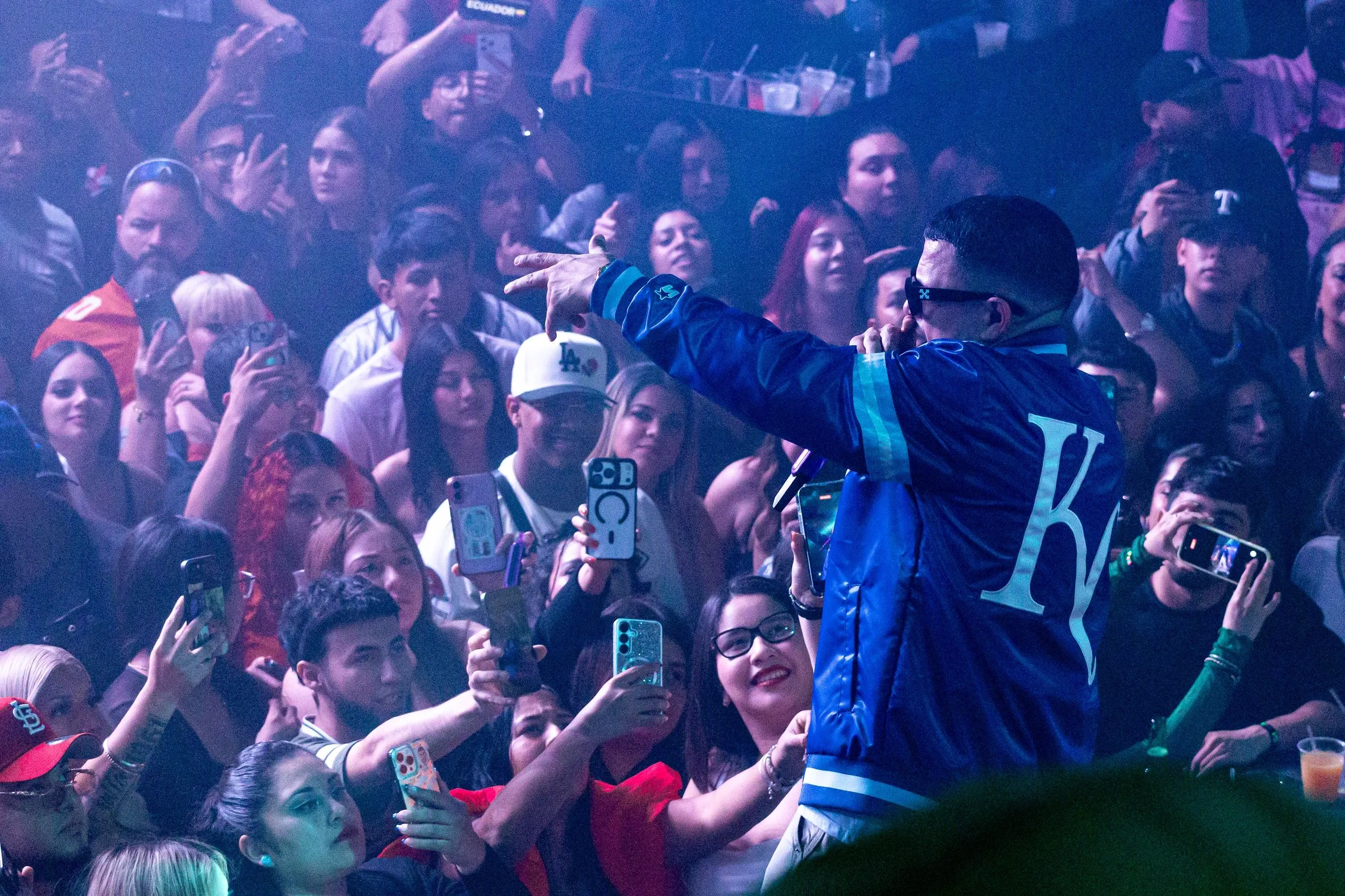 An artist in a blue satin baseball jacket leaning out over a wall of raised phones.