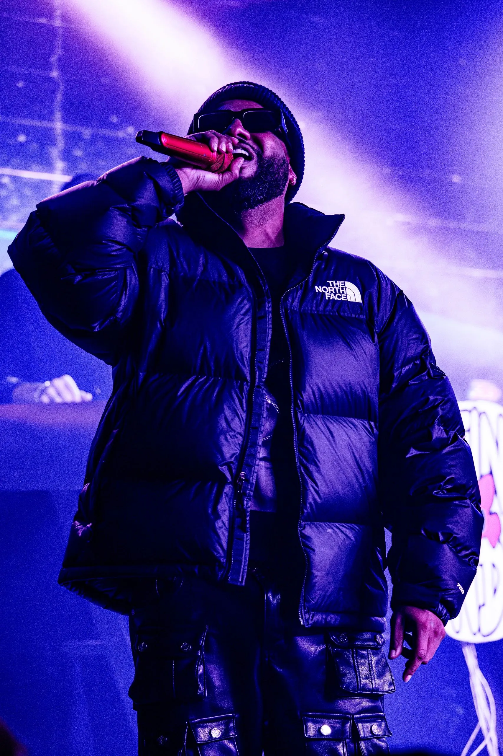 An artist on the microphone in a black puffer jacket, lit from behind in purple.