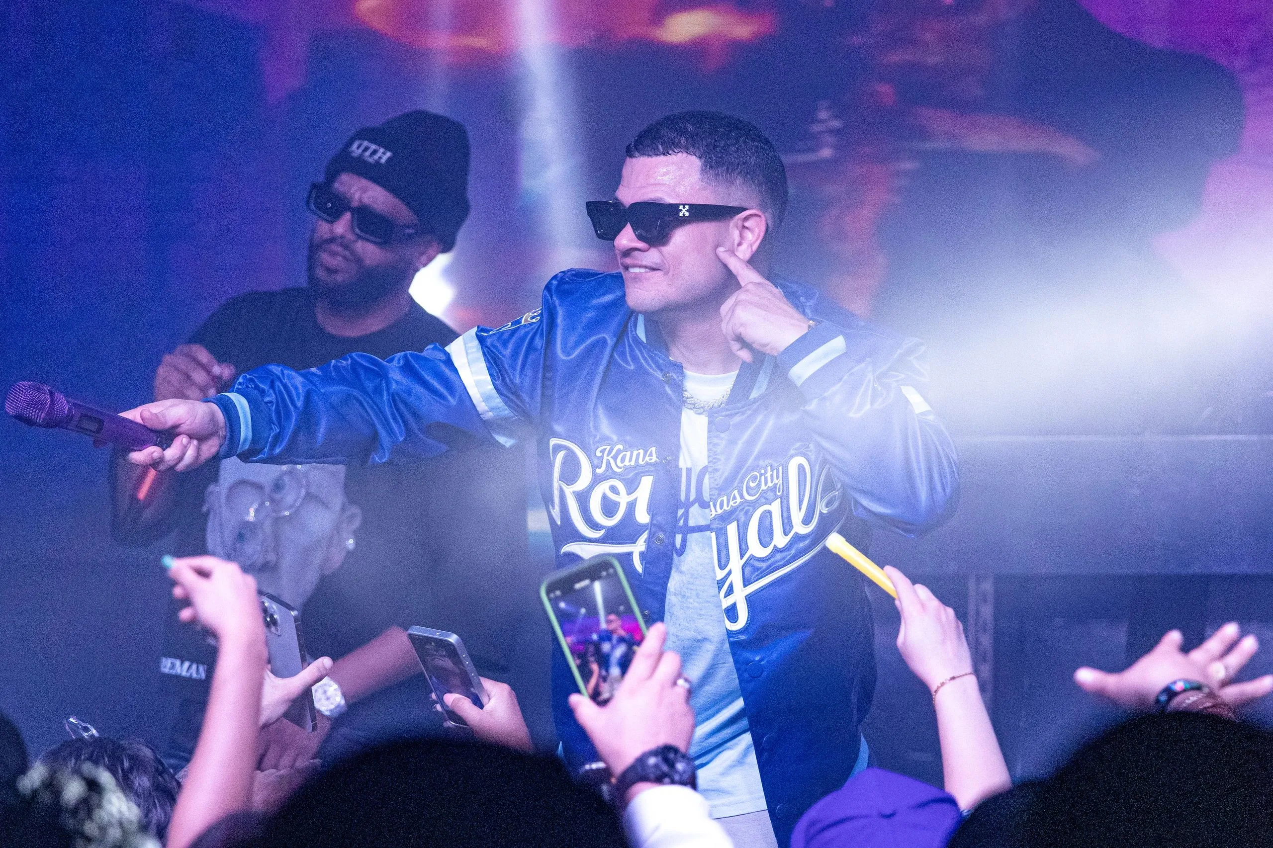 An artist in a blue satin baseball jacket pointing into the crowd while phones film from the front row.