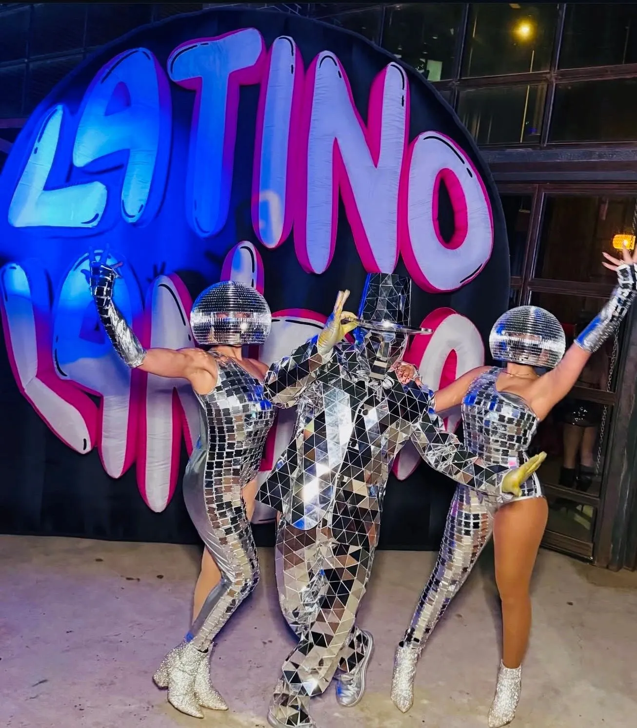 Three performers in mirror-tile bodysuits and disco-ball helmets posing in front of an inflated Latino Landia bubble wordmark.